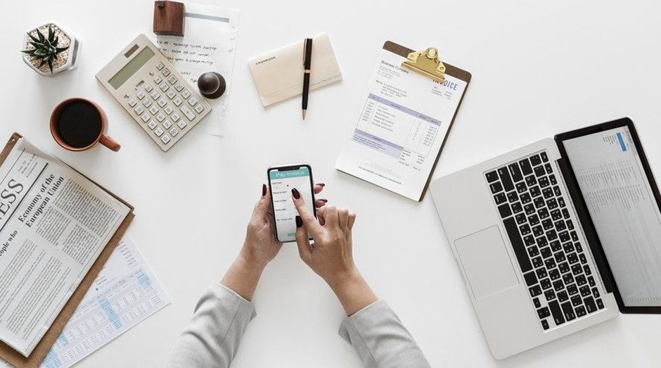Mesa de trabalho com notebook, celular, jornal e materiais de escritório